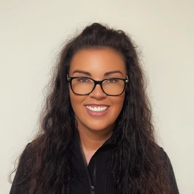Dental Assistant April In Black Top Smiling In Front Of Beige Wall