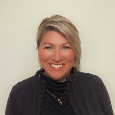 Patient Care Coordinator Shelly In Black Top Smiling In Front Of Beige Wall